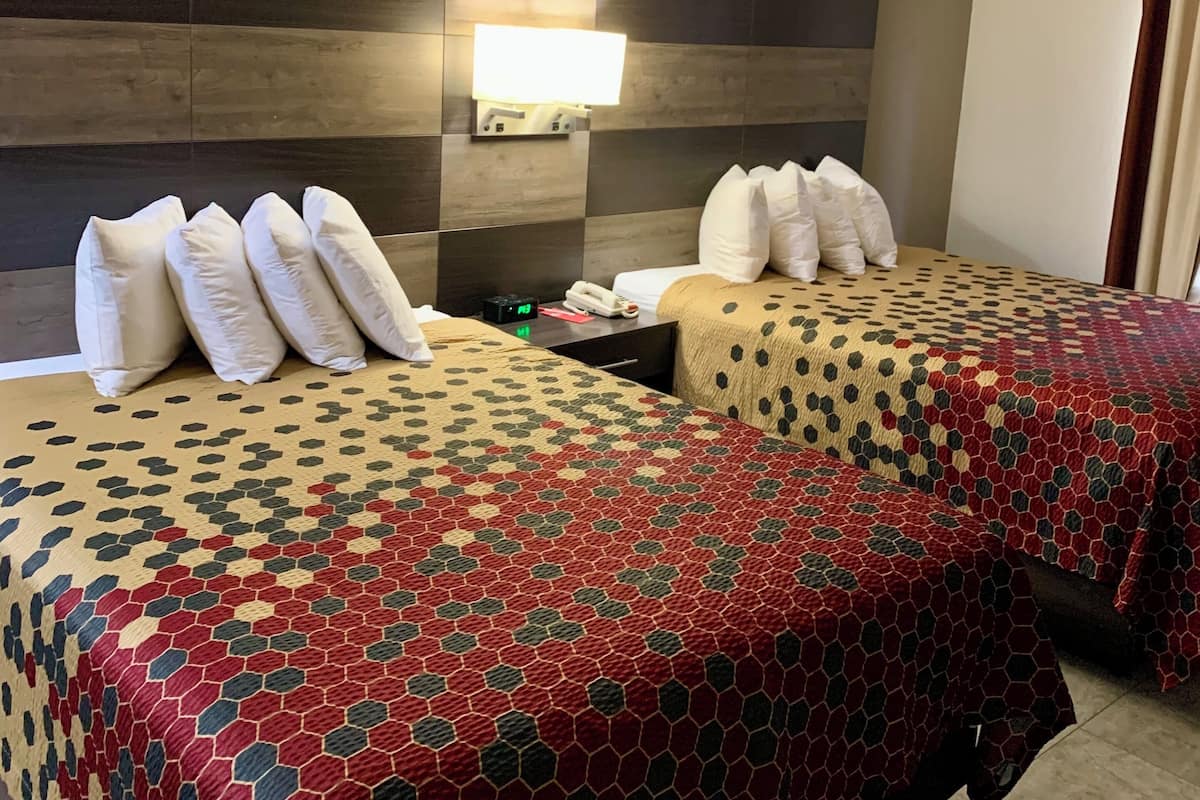 A hotel room with two beds, each with a patterned bedspread, a wall-mounted light, and a nightstand with a phone and a small table.