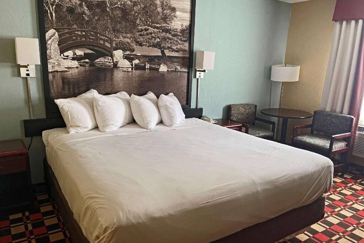 A hotel room with a large bed, two bedside tables, a chair, a small table, and a large framed picture on the wall.