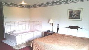 Quarto de hotel com uma cama grande, banheiro separado com banheira, mesa de cabeceira com abajur e um quadro na parede.