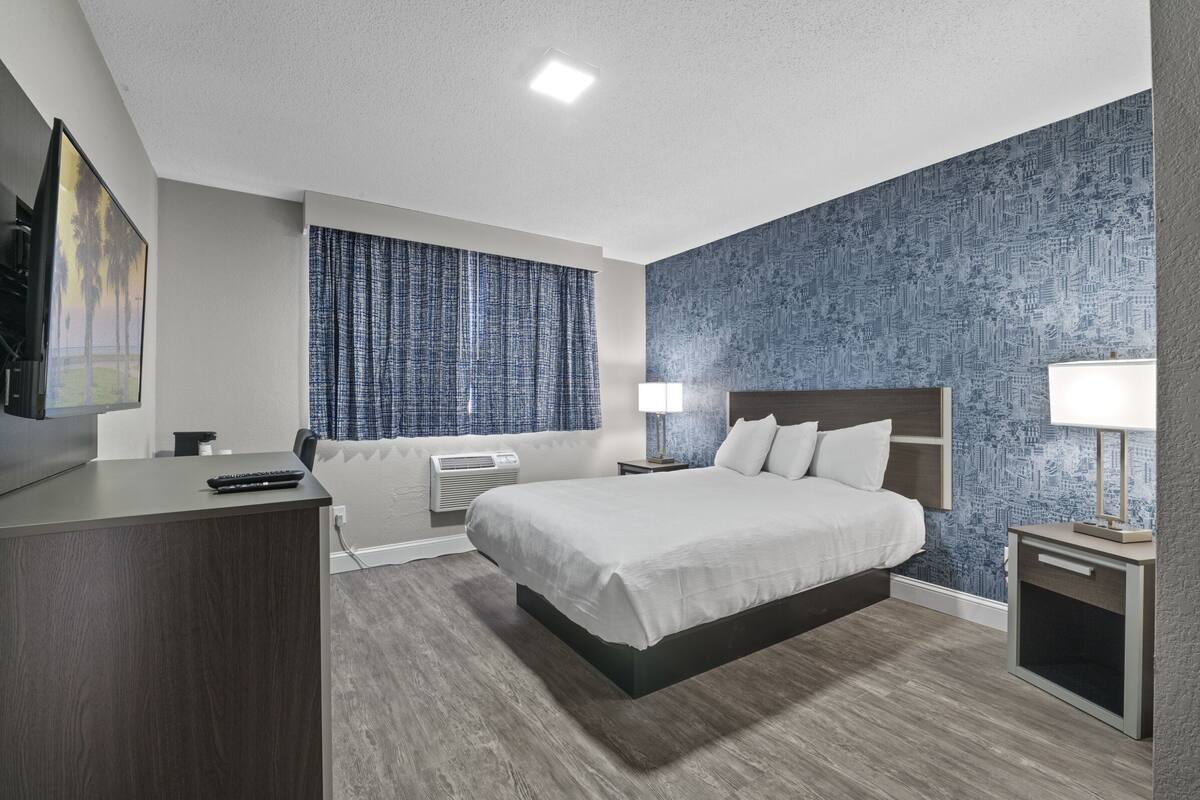 A modern hotel room with a large bed, a desk, a chair, a television, and a window with curtains.