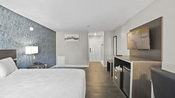 A hotel room with a large bed, a desk with a chair, a TV mounted on the wall, and a cityscape artwork on the wall.