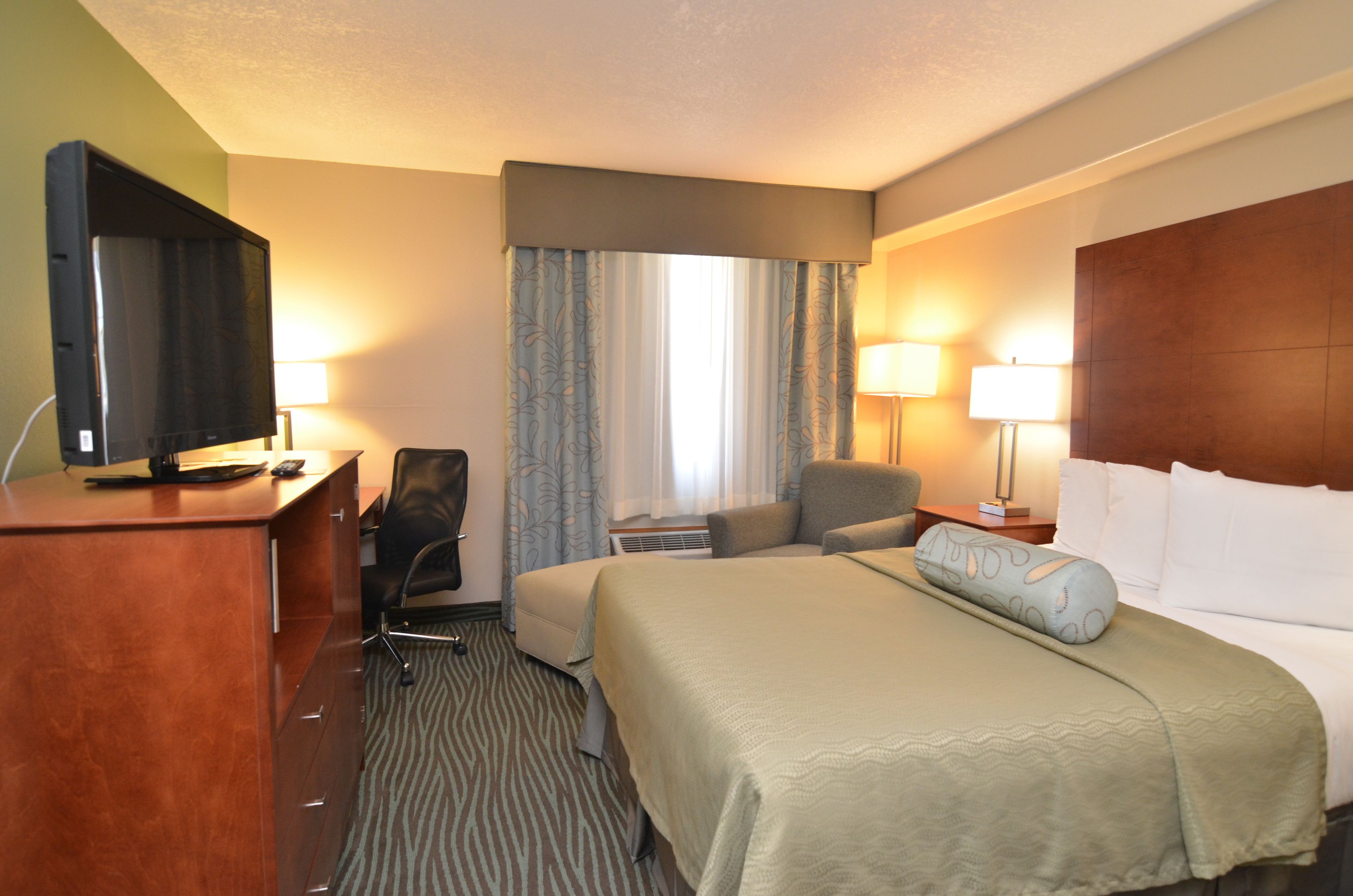 A hotel room with a bed, a desk with a television, a chair, a lamp, and a window with curtains.