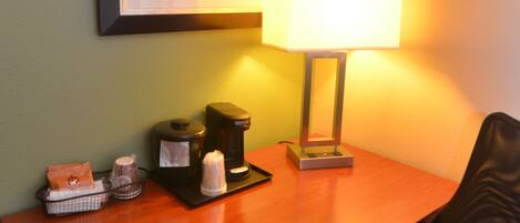 A hotel room with a wooden desk, a lamp, a coffee maker, and a framed picture on the wall.