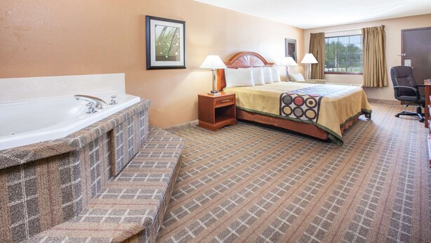 A hotel room with a large bathtub, a bed with a patterned bedspread, two bedside lamps, and a chair. - Super 8 by Wyndham Clinton (Clinton)