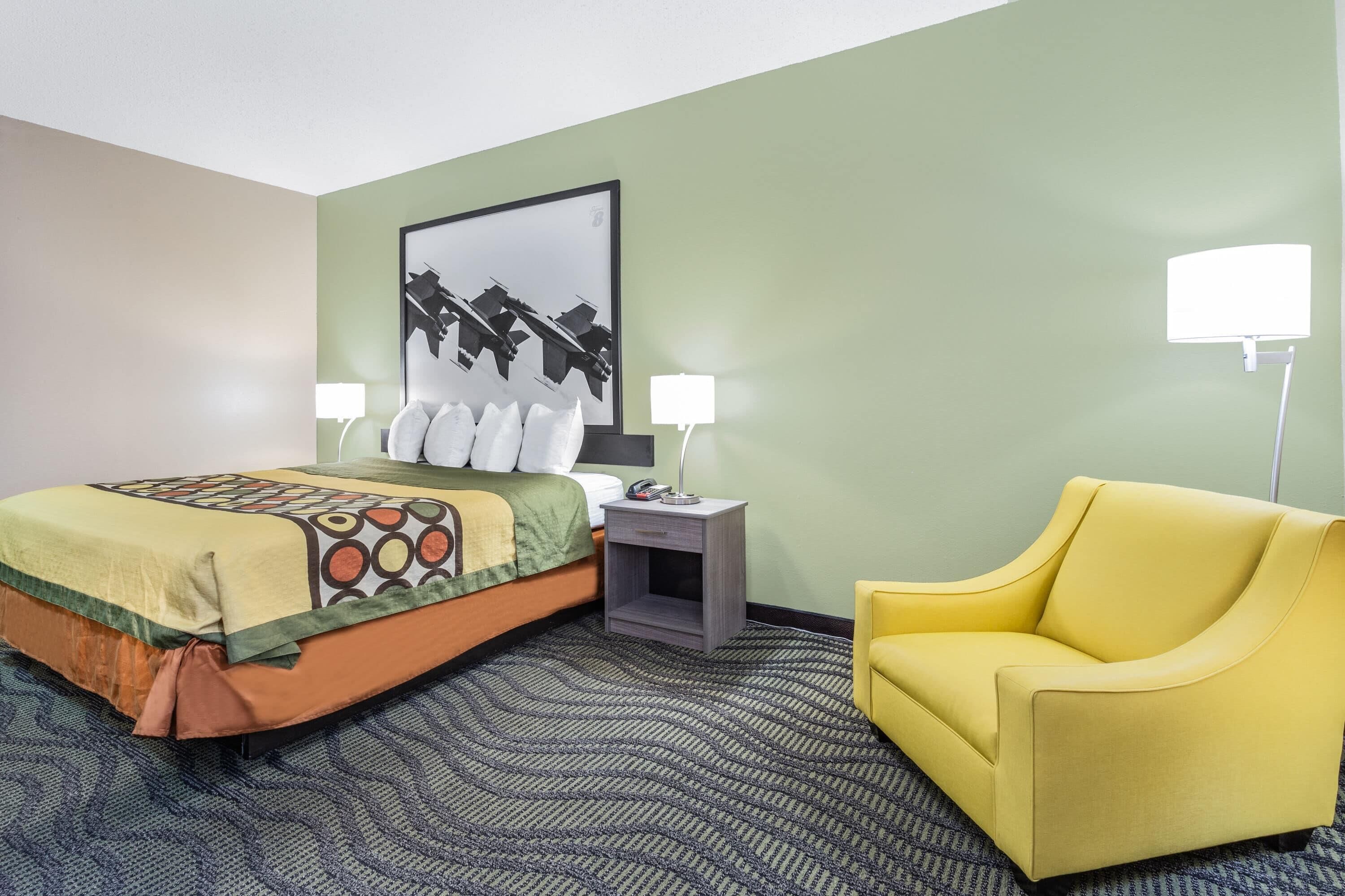 A hotel room with a bed, a yellow armchair, a nightstand with a lamp, and a framed picture on the wall.