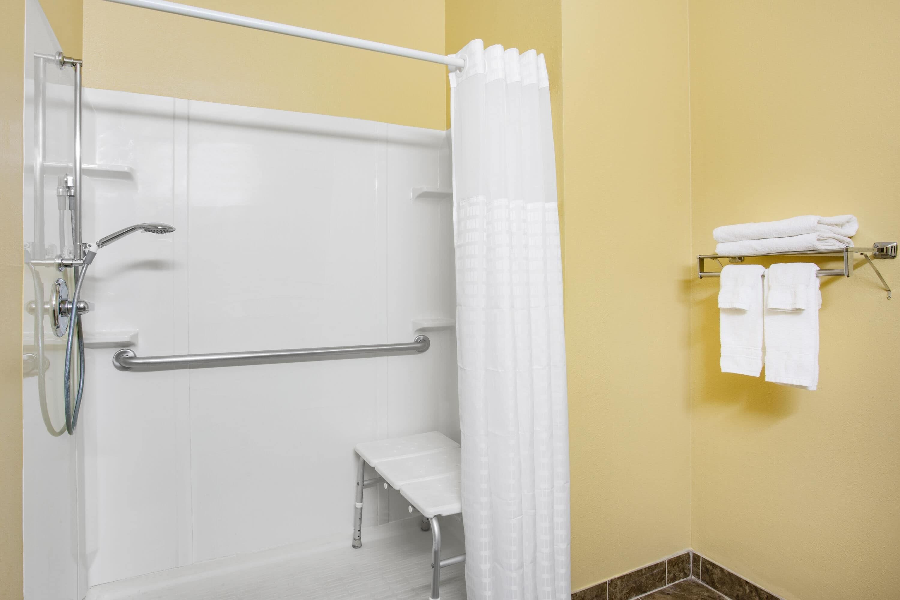 A shower with a white curtain, a grab bar, and a bench. There are towels on a rack.