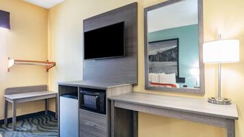 Habitación de hotel con televisión de pantalla plana montada en la pared, un refrigerador pequeño, un microondas, un escritorio con cajones, un banco y una lámpara.