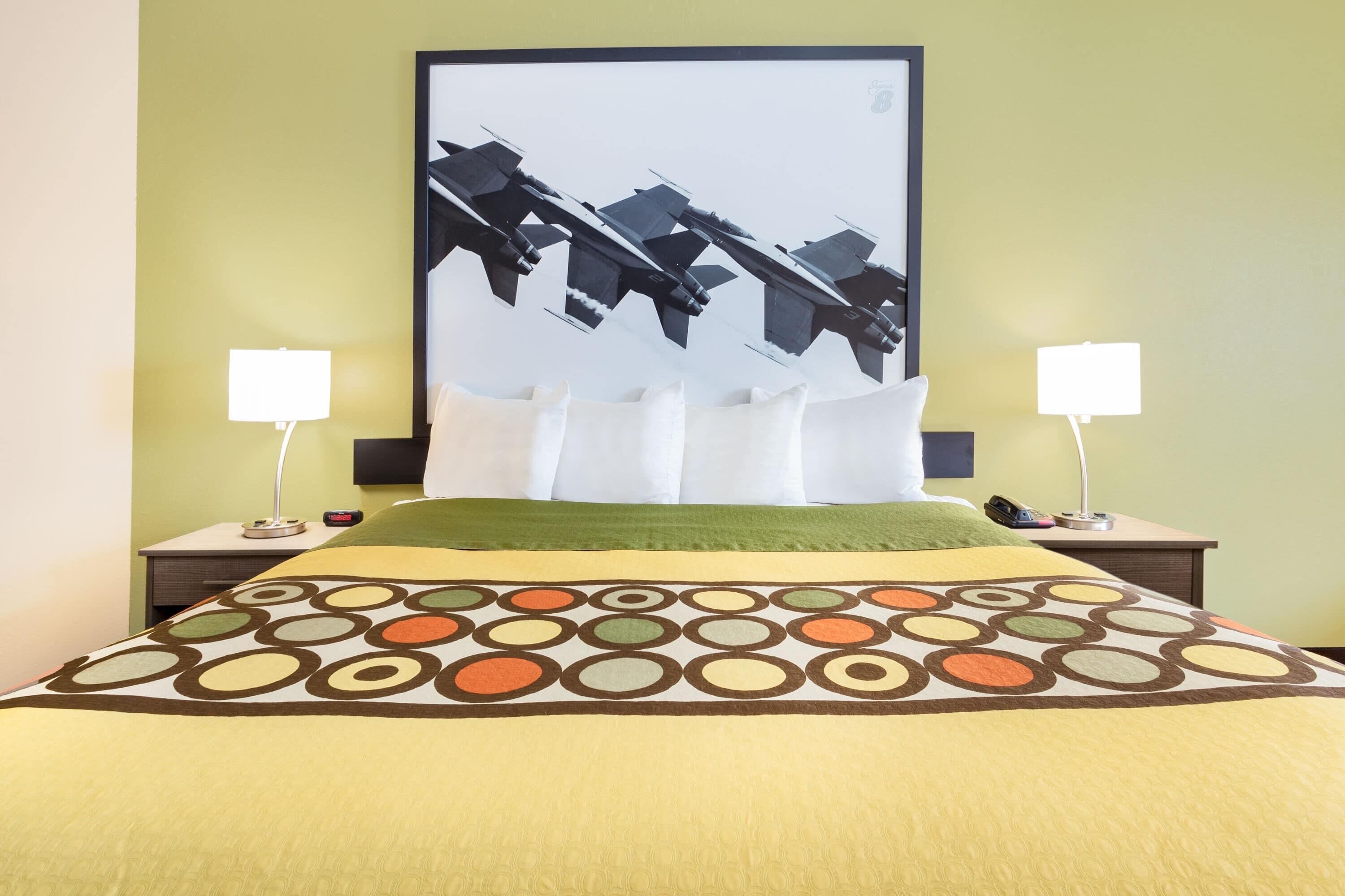 A hotel room with a bed, green walls, a large framed picture of fighter jets, two bedside tables with lamps, and a patterned bedspread.