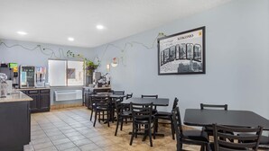 Un restaurante con piso de baldosas, mesas y sillas de comedor negras, una zona de cocina con refrigerador, y un letrero en la pared que dice 'Greetings From Kokomo'.