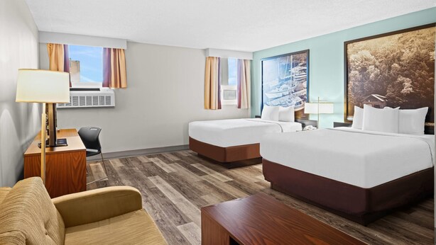 A hotel room with a large bed, a smaller bed, a desk, a chair, and a couch. There is a window with curtains, a wall-mounted air conditioner, and a painting on the wall.