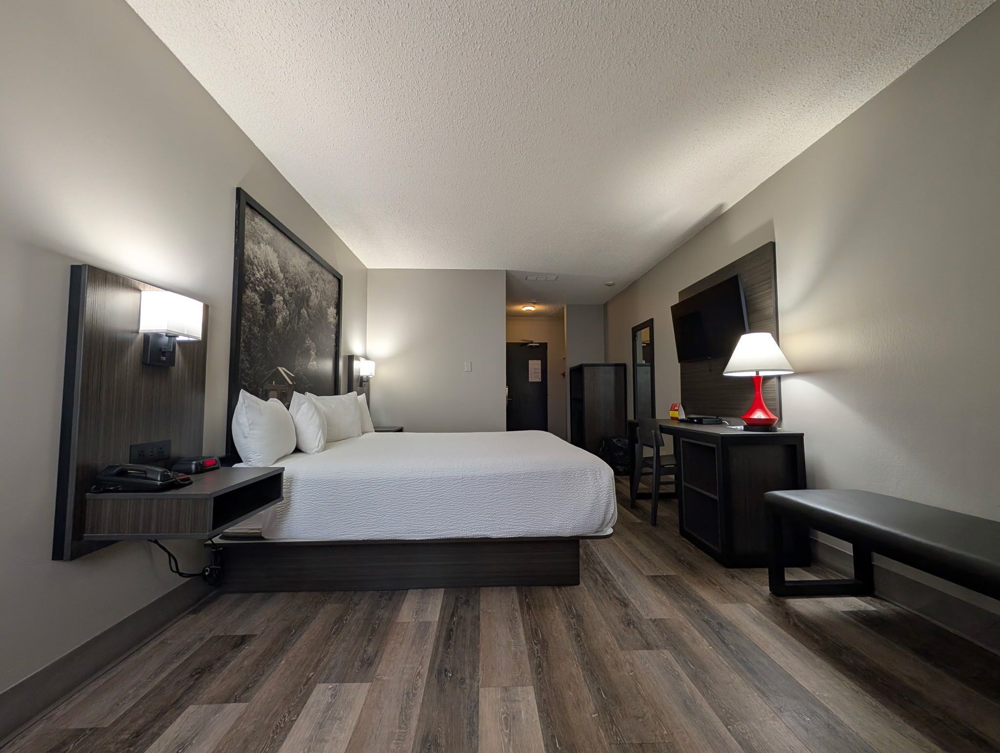 A hotel room with a large bed, a desk with a lamp, a TV, and a bench.
