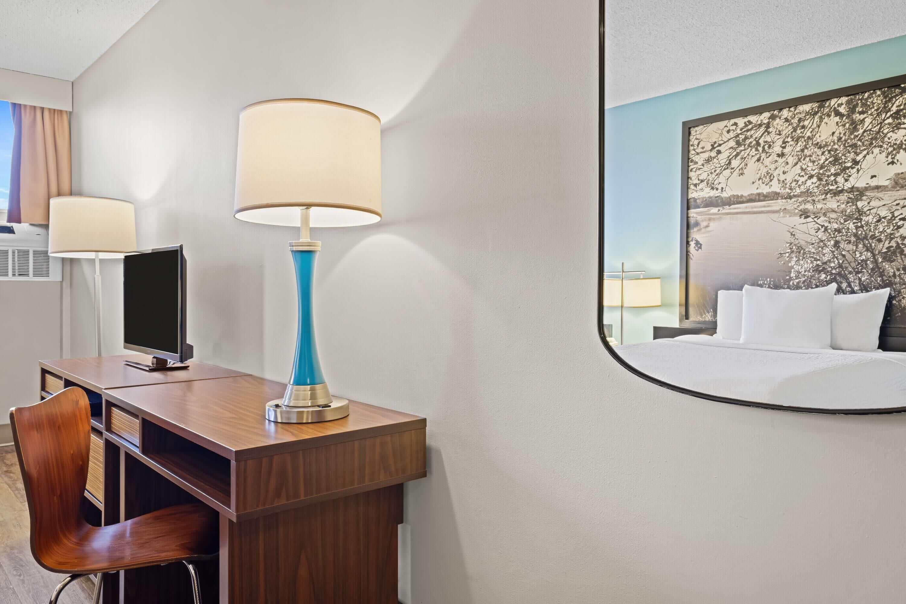 A hotel room with a wooden desk, two lamps, a television, and a large framed picture of a landscape.