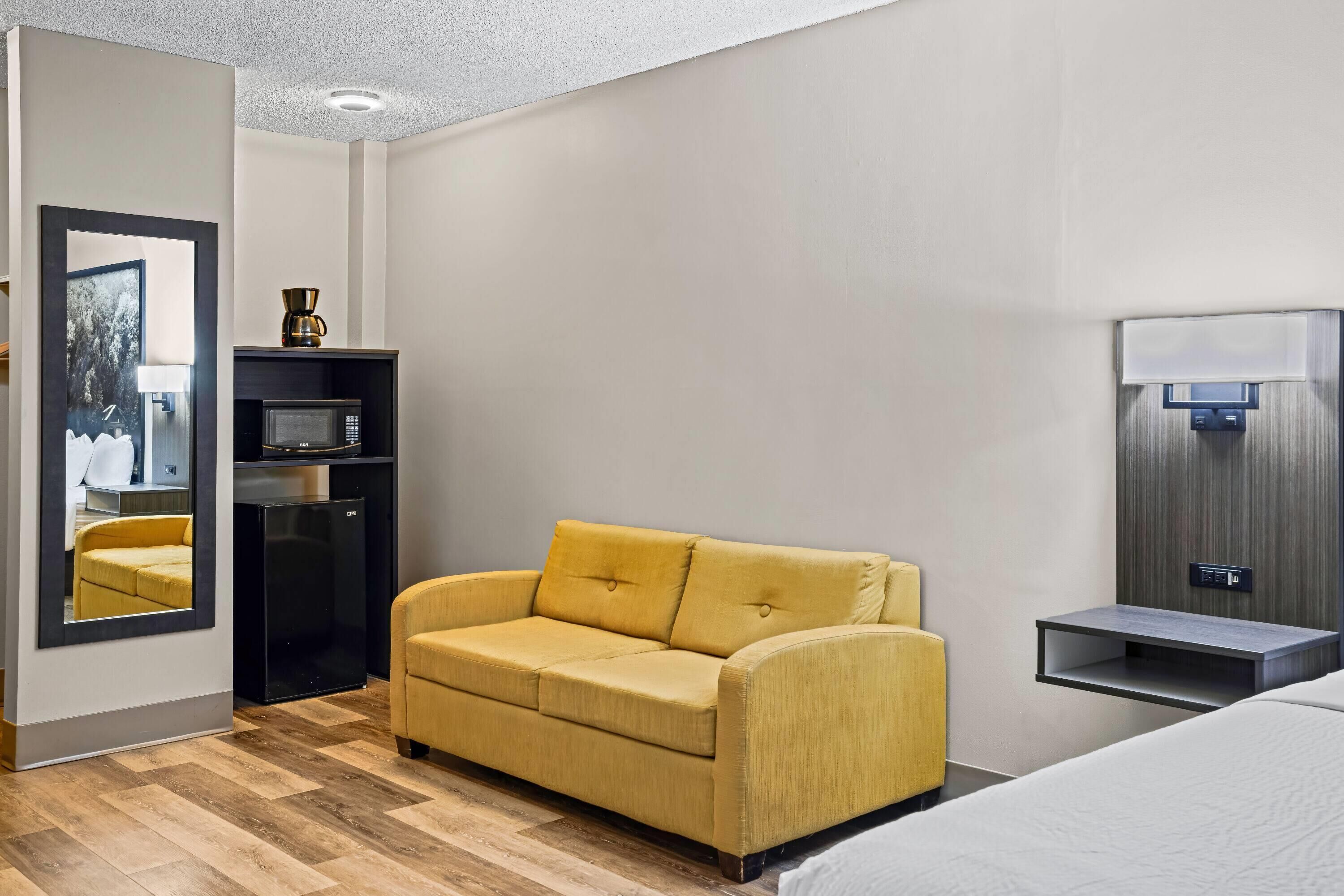 Ein Hotelzimmer mit gelber Couch, einem kleinen schwarzen Kühlschrank, einer Mikrowelle, einem Nachttisch und einem Spiegel.