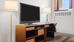 A hotel room with a flat-screen TV on a wooden TV stand, two wall-mounted lamps, a window with curtains, and an air conditioning unit.