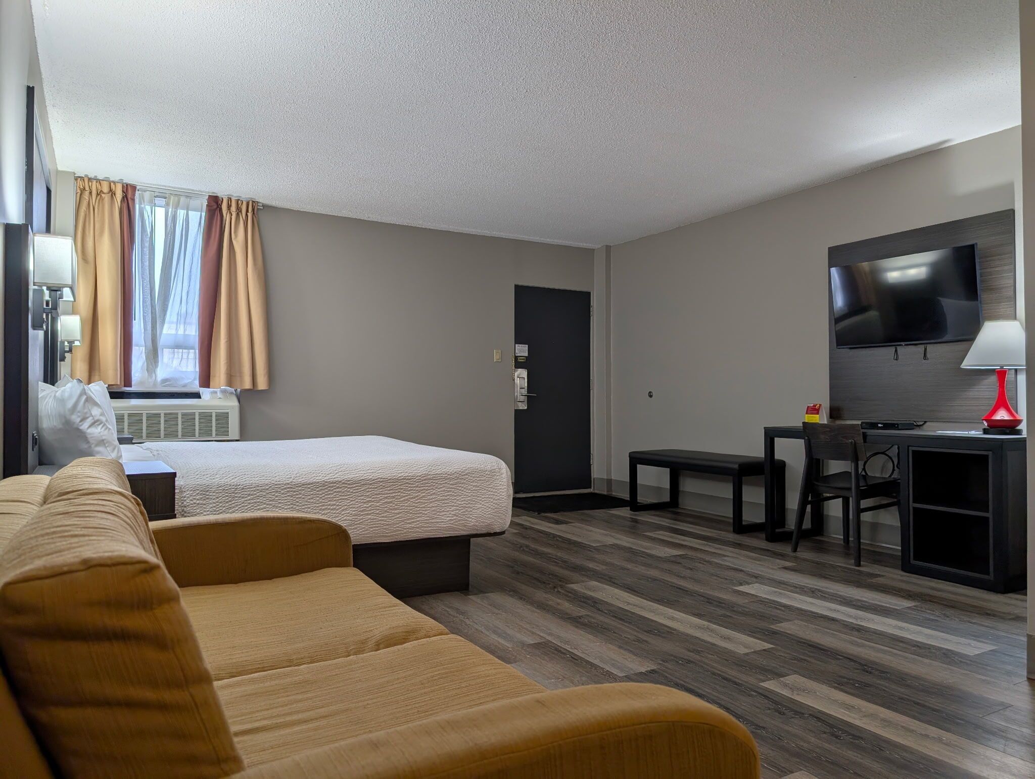 A hotel room with a bed, a sofa, a desk with a chair, a TV, and a window with curtains.