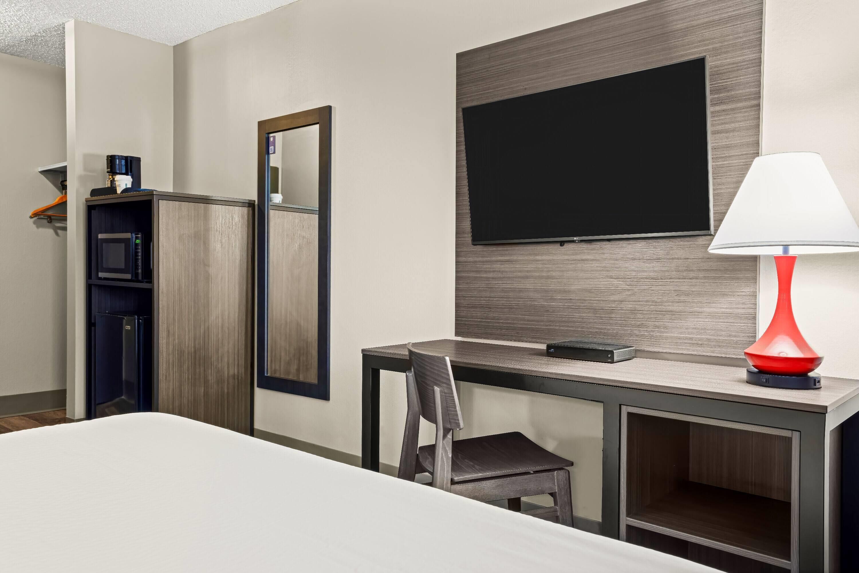 A hotel room with a bed, a desk with a chair, a TV mounted on the wall, a red lamp, and a microwave.