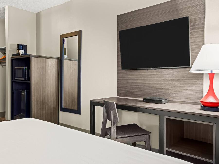 A hotel room with a bed, a desk with a chair, a TV mounted on the wall, a red lamp, and a microwave.