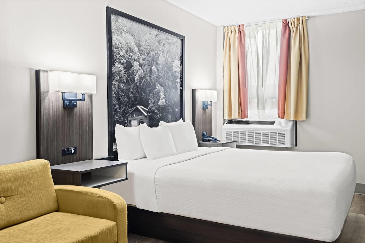 Une chambre d’hôtel avec un grand lit, deux tables de chevet, une petite table, un fauteuil jaune, un grand tableau encadré et une fenêtre avec des rideaux.