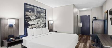 Habitación de hotel con una cama grande, un escritorio, una silla, una lámpara de escritorio, una mesita de noche, un teléfono montado en la pared y un cuadro con barcos.