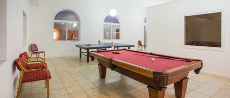 Ein Billardtisch mit roter Filzauflage, zwei Billardstöcke und ein roter Sessel. Zwei Fenster mit Rundbögen, eines davon mit Einblick ins Innere.