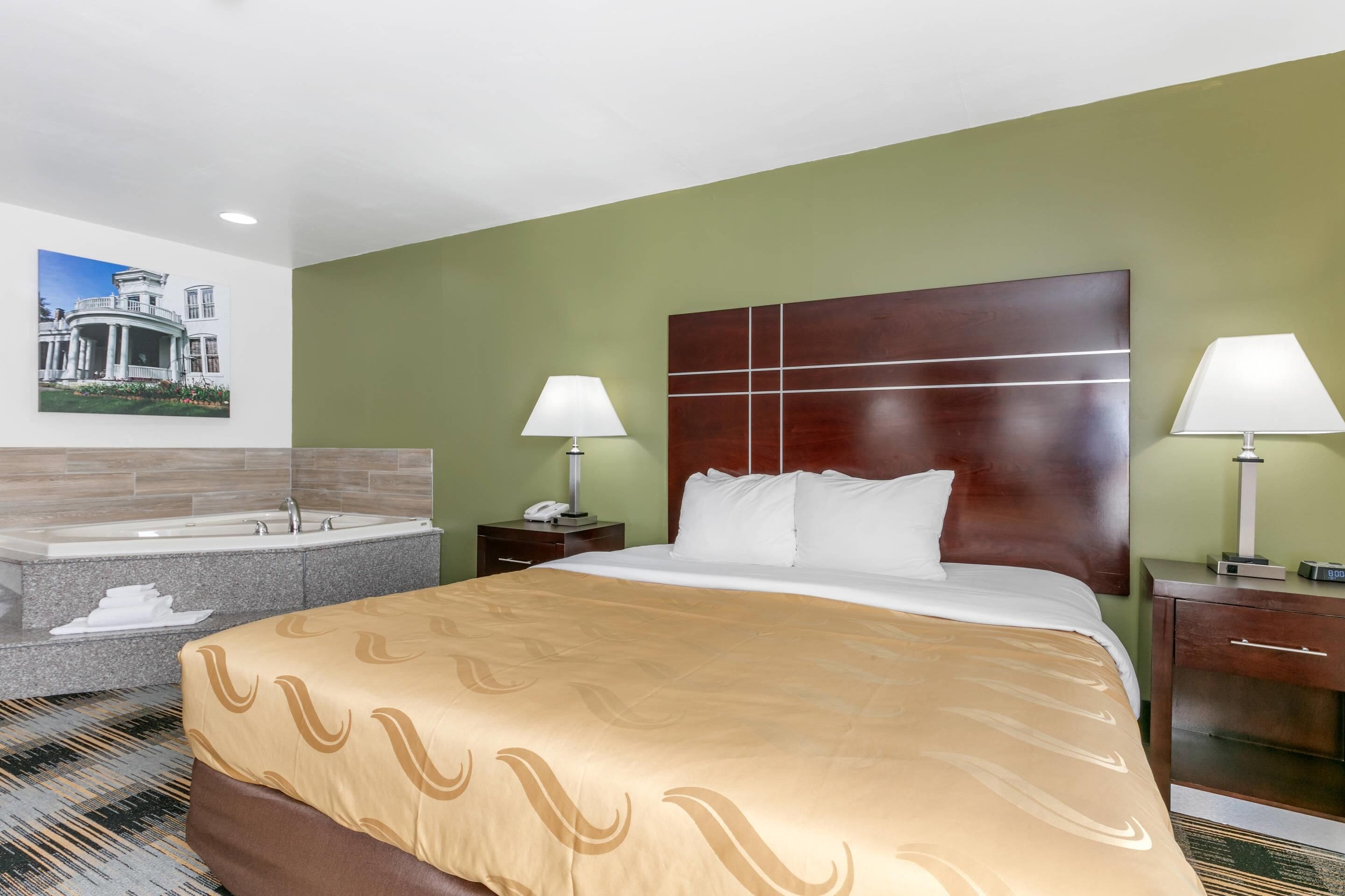 Habitación de hotel con una cama grande, dos mesitas de noche con lámparas, una bañera integrada y un cuadro colgado en la pared.