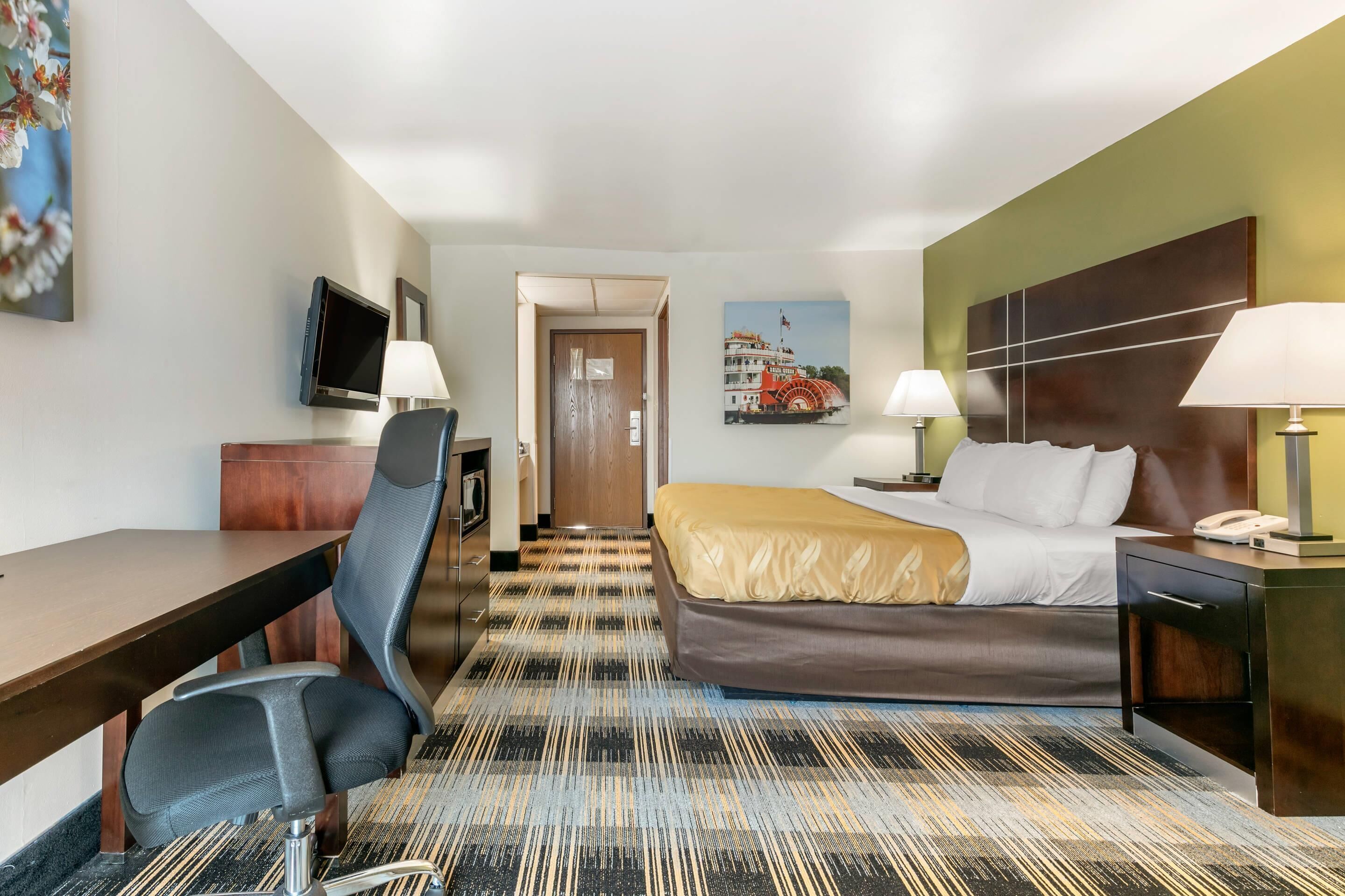 A hotel room with a bed, desk, chair, TV, lamps, and a painting on the wall.