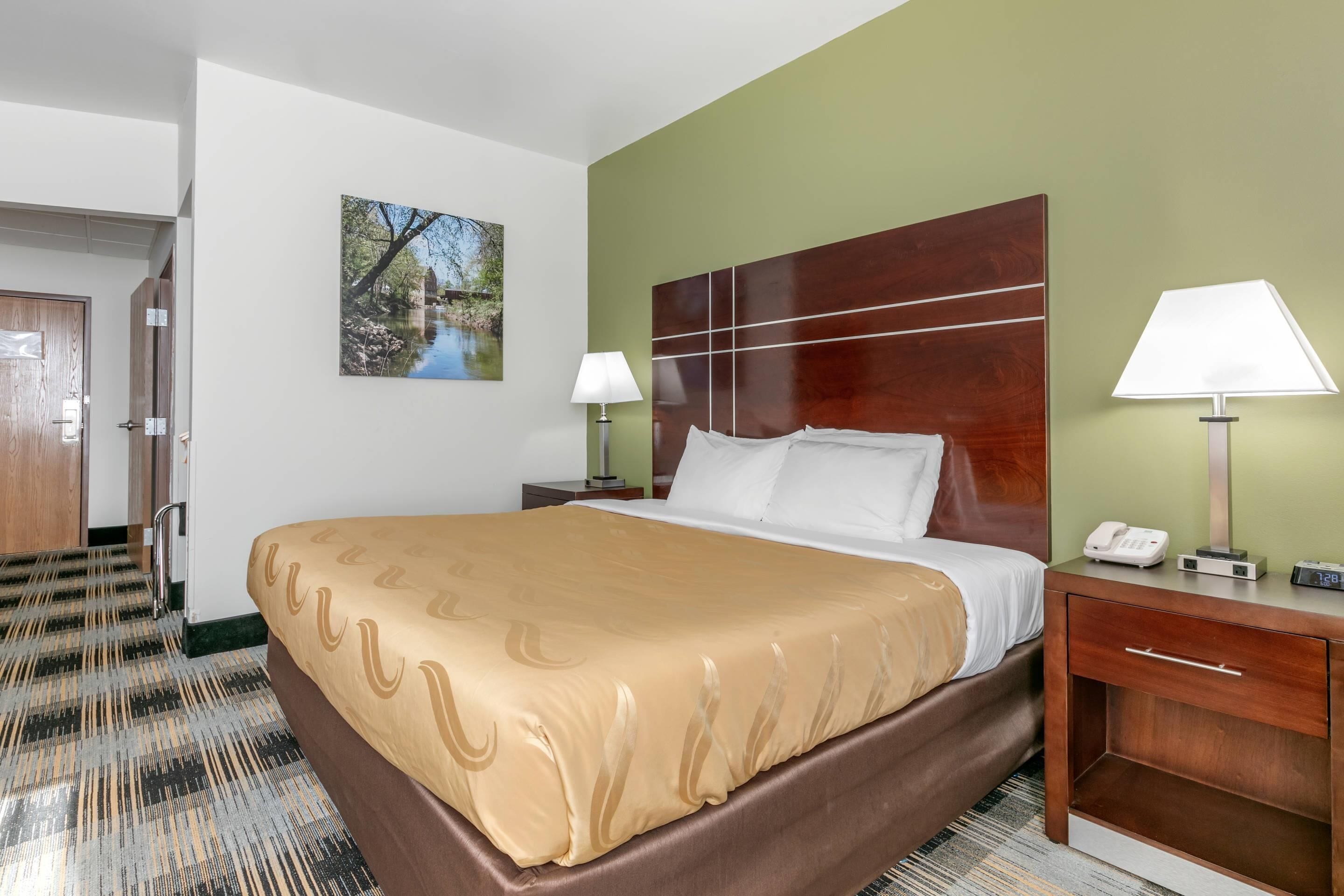 A hotel room with a large bed, two bedside lamps, a wooden headboard, a painting on the wall, and a door leading to another room.