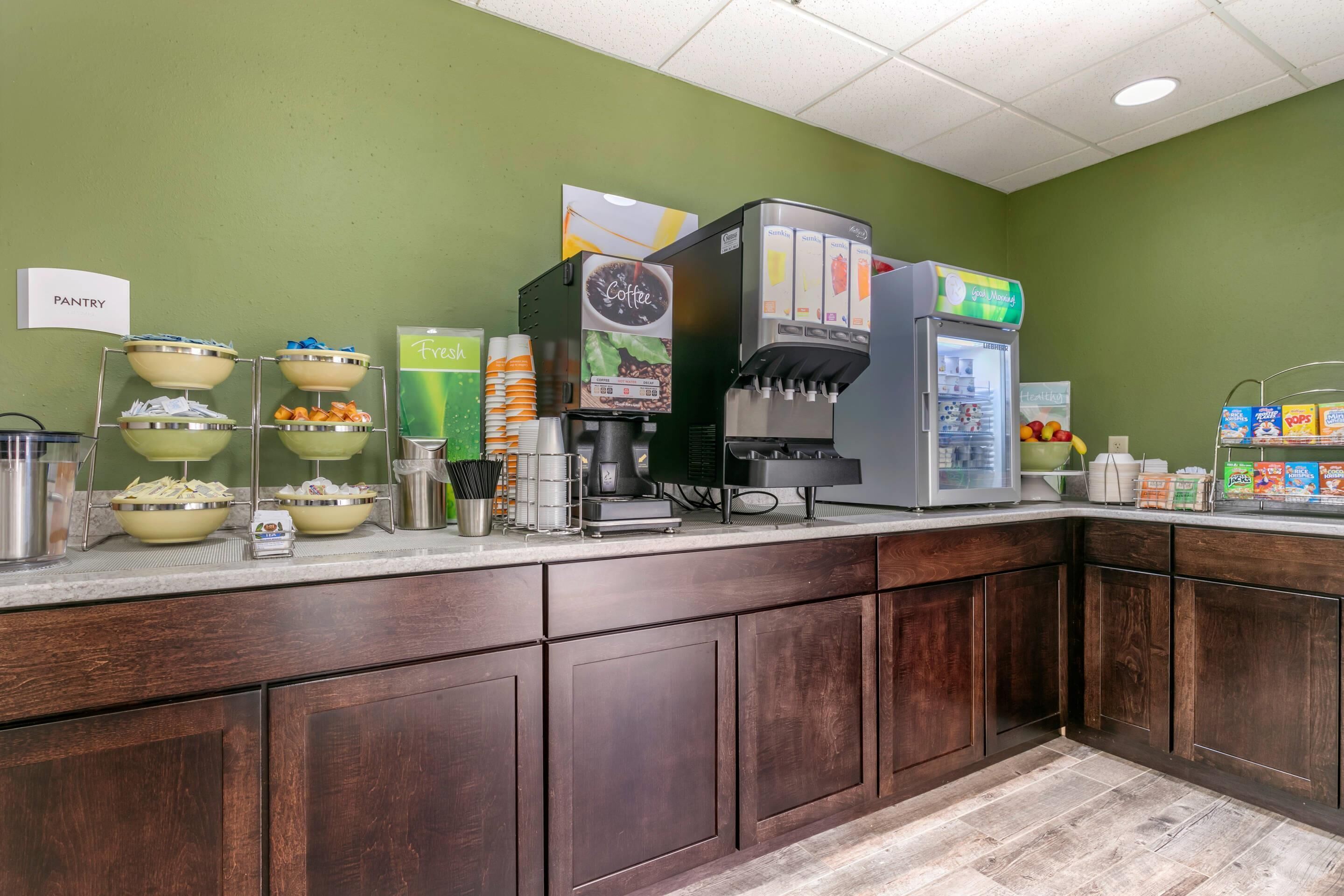 A self-service breakfast station with coffee machines, a refrigerator, and various food items.