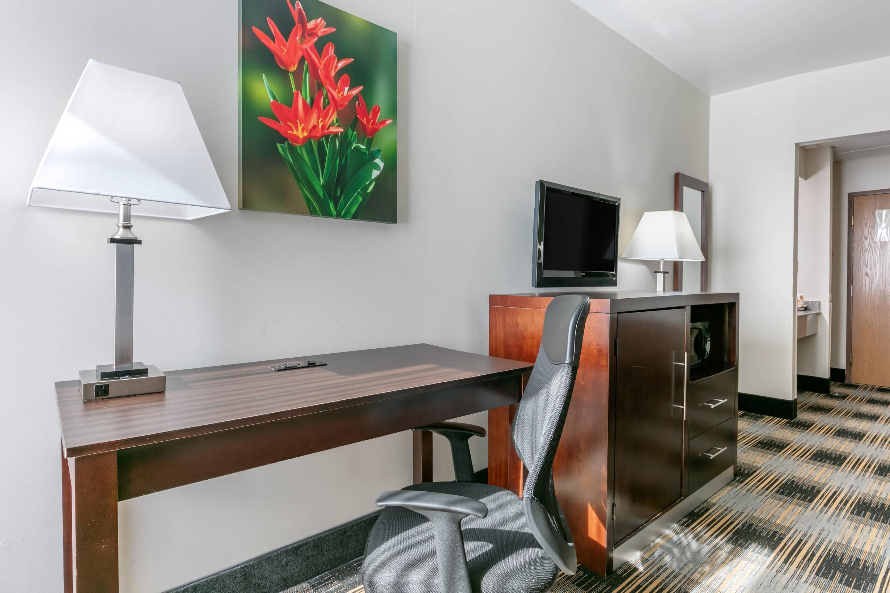 A hotel room with a desk, chair, TV, and a painting of flowers.