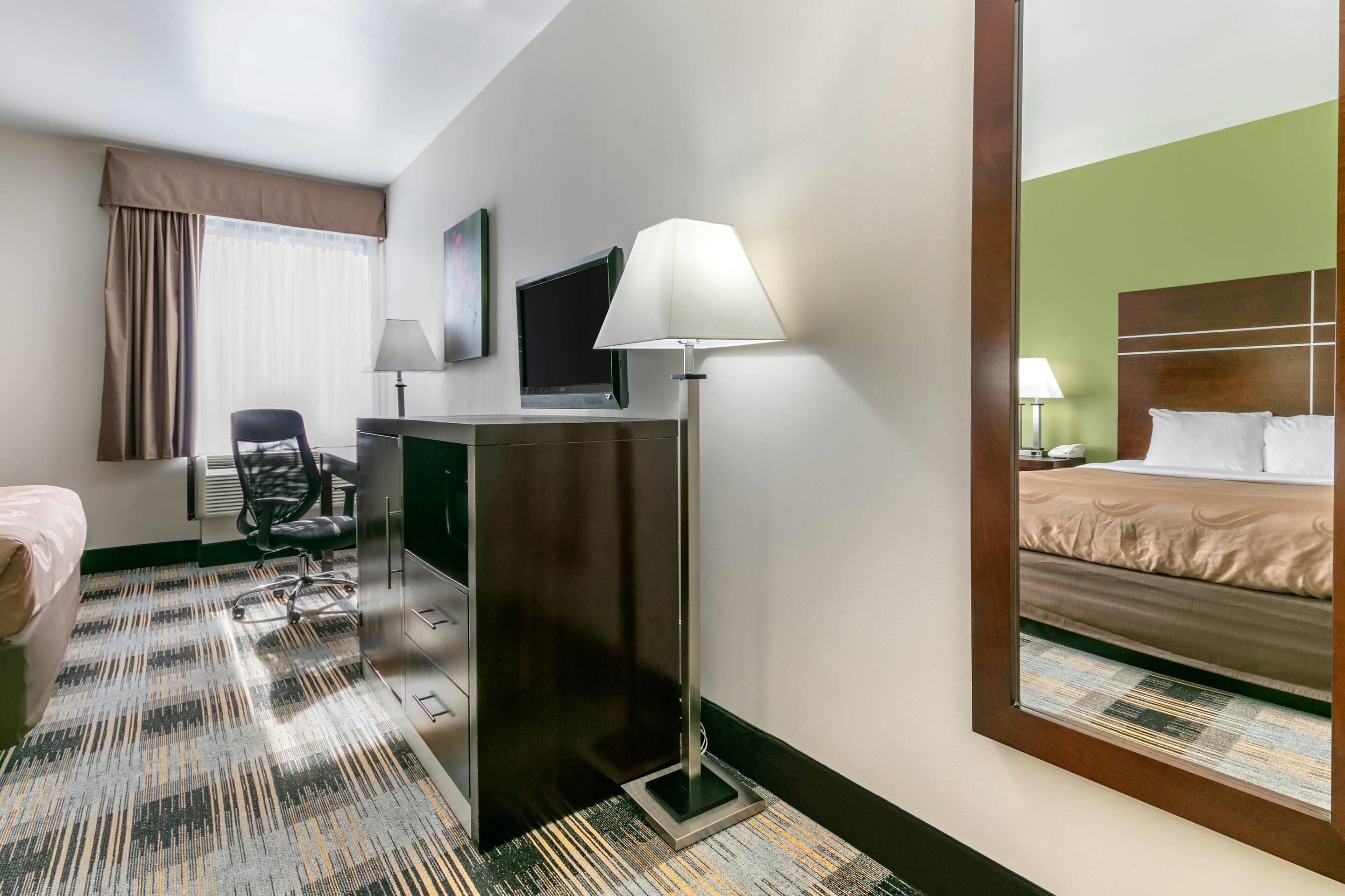 Habitación de hotel con cama, escritorio, silla, lámpara y un espejo grande que refleja la cama.