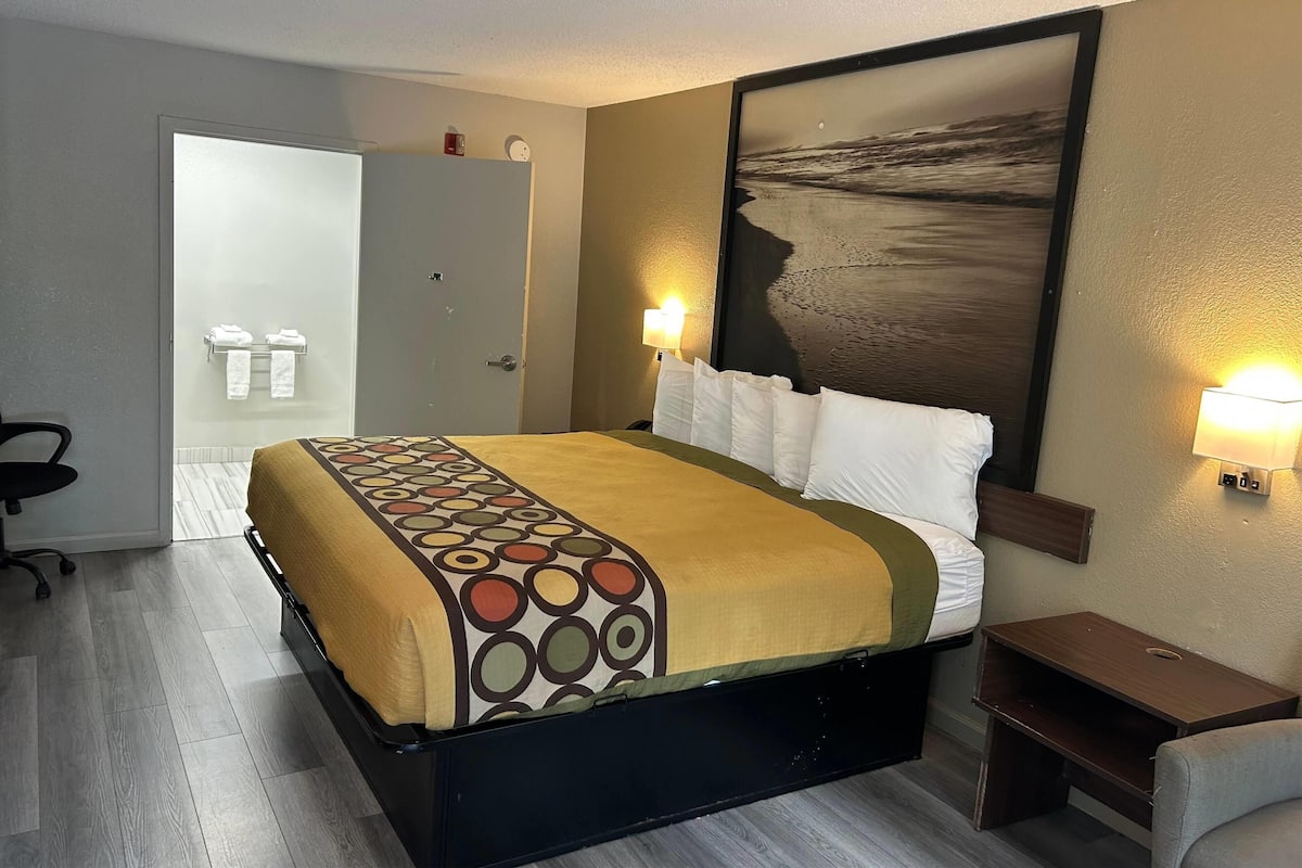 Una habitación de hotel con cama, escritorio, silla, mesita de noche, lámpara de pared y puerta que da al baño.