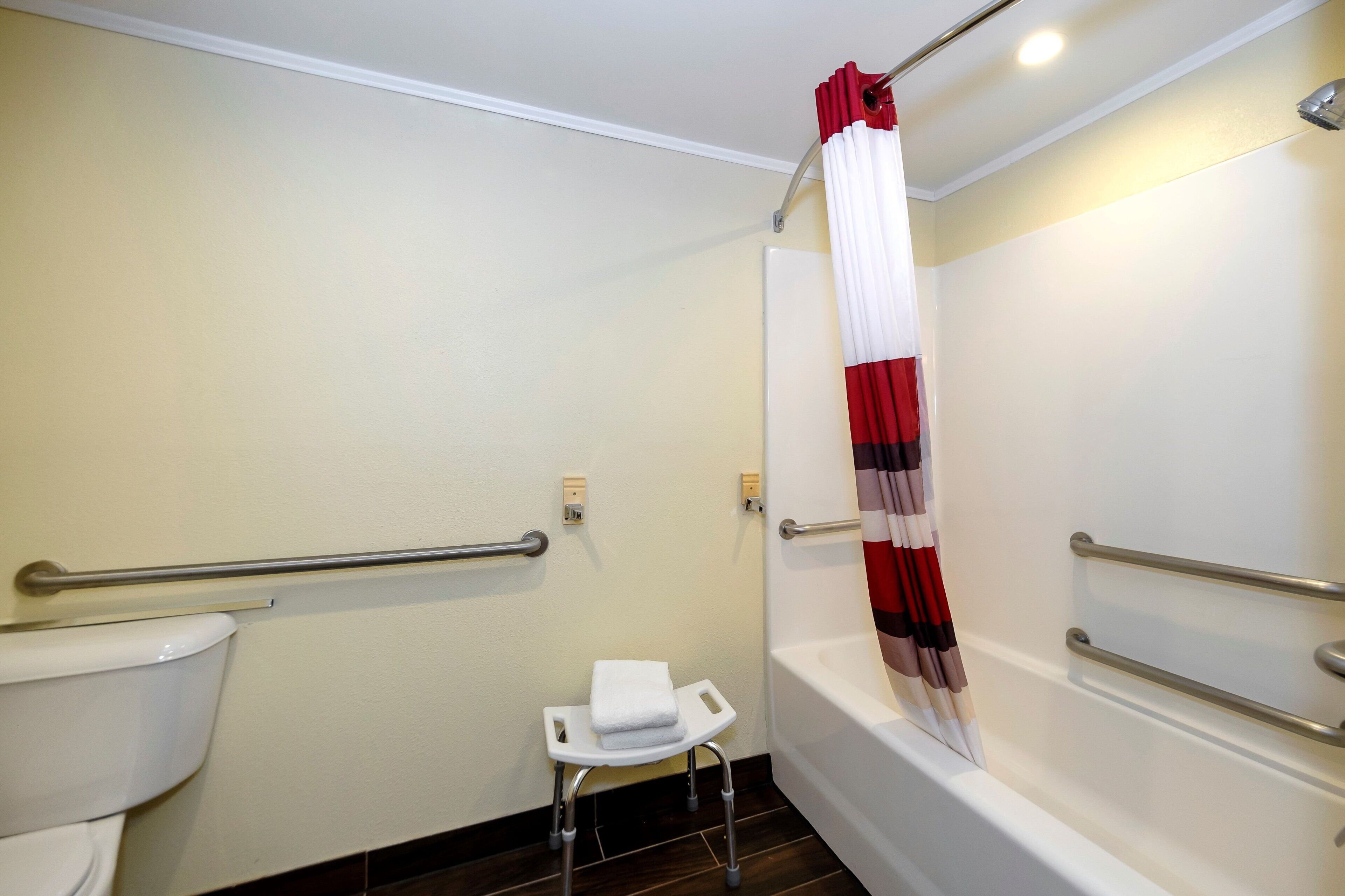 Un baño con inodoro, ducha con cortina roja y blanca, y un taburete pequeño y blanco.