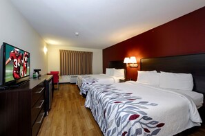 A hotel room with a television, a desk with a coffee maker, a bed with a floral bedspread, a bedside table with a lamp, and a window with curtains.