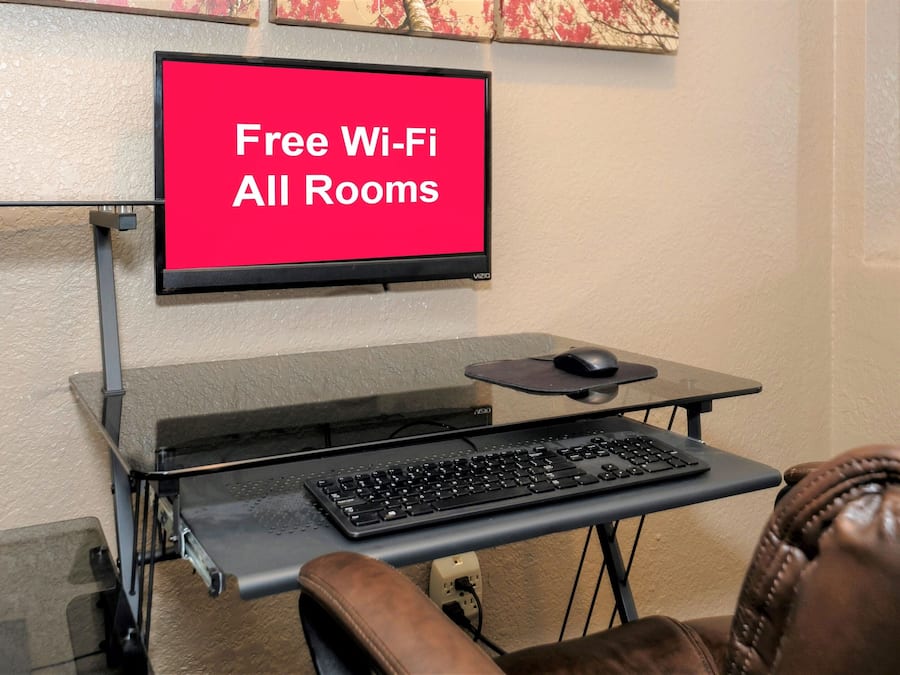 Escritorio con teclado y ratón, un monitor mostrando 'Free Wi-Fi All Rooms' y una estantería de pared.