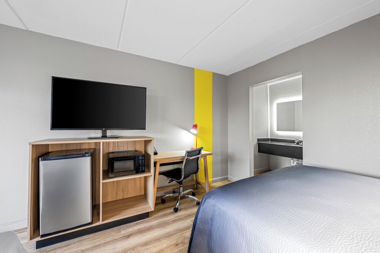 Una camera d'albergo con un letto, una scrivania con un computer, una televisione, un frigorifero e un bagno con uno specchio.