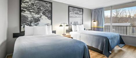 A hotel room with two beds, a large window with a view, and artwork on the walls.