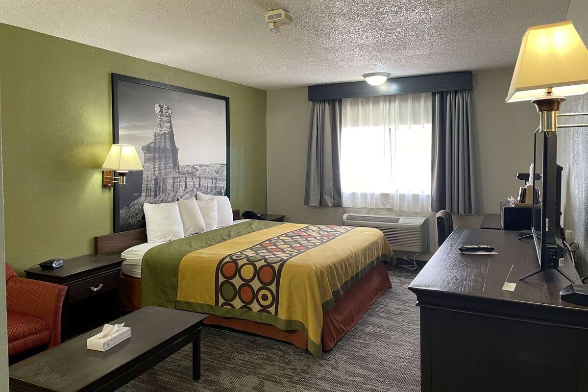 A hotel room with a bed, a desk, a chair, a lamp, a window with curtains, and a wall-mounted air conditioner.