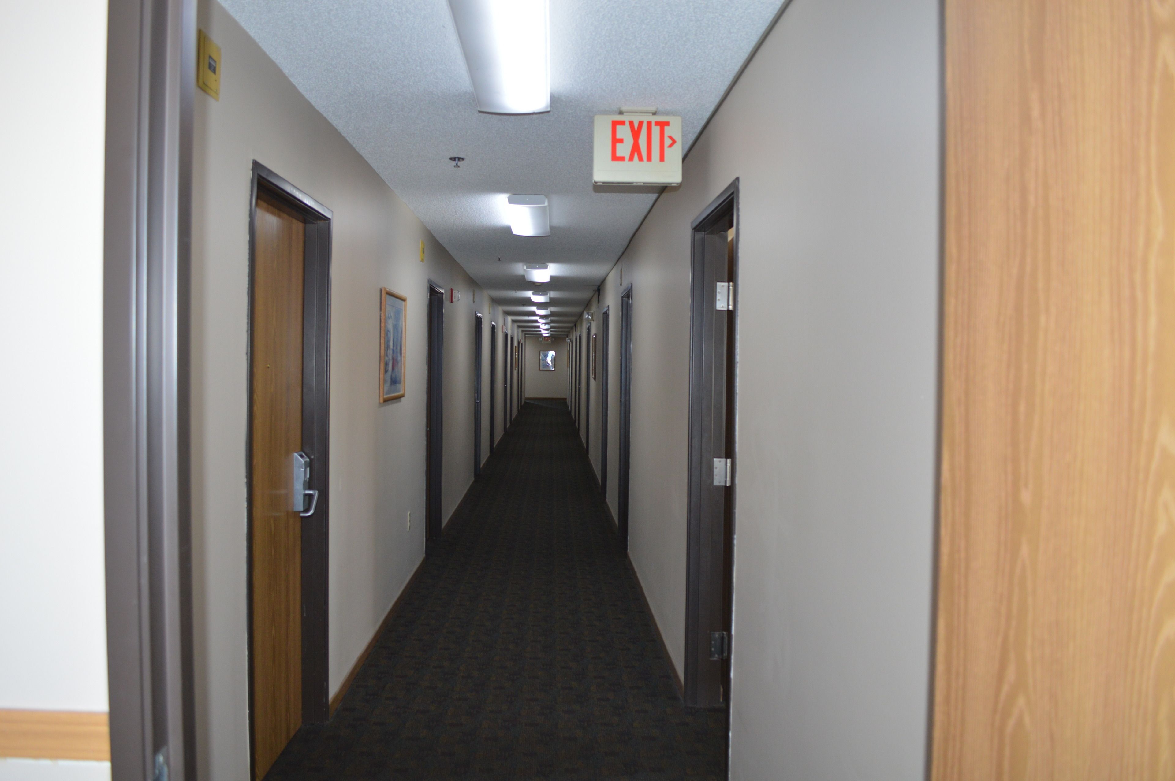 A long, narrow hotel corridor with doors on both sides, an EXIT sign above, and a carpeted floor.