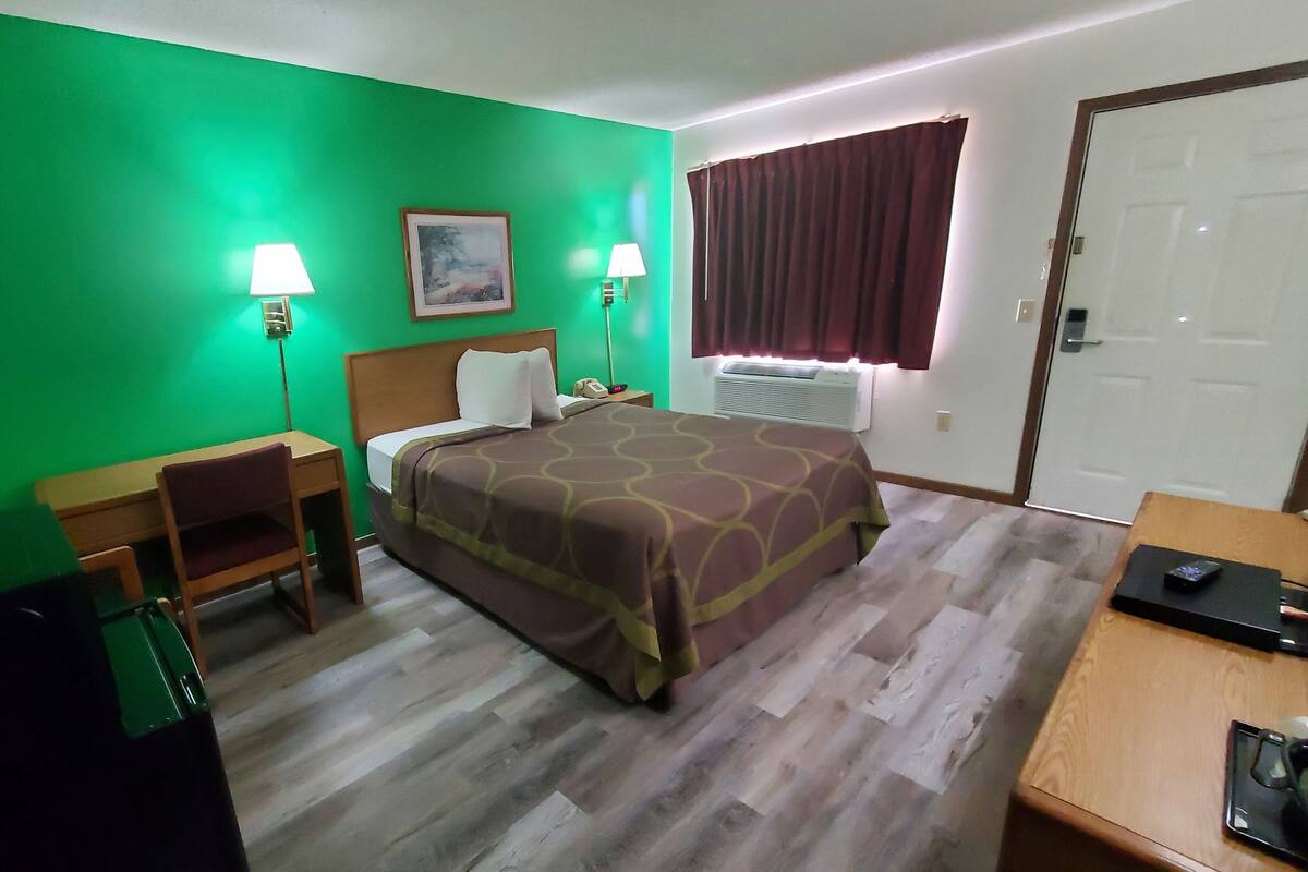 Habitación de hotel con cama, escritorio, silla, dos lámparas de noche, un cuadro en la pared, ventana con cortinas y una puerta.