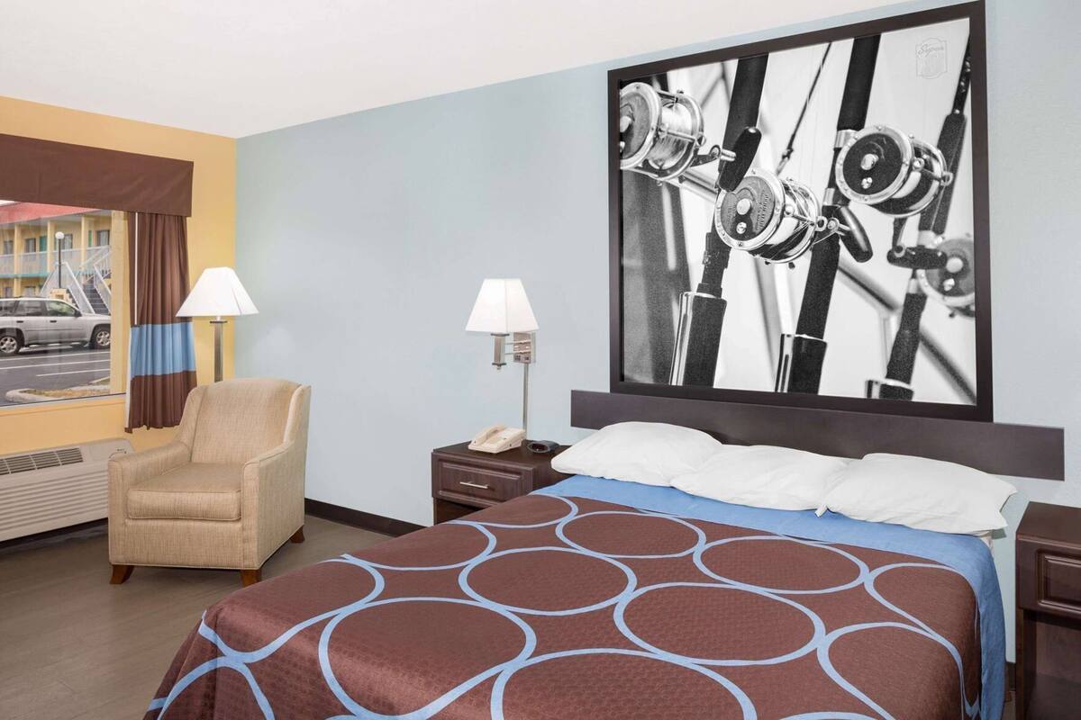 Une chambre d’hôtel comprenant un lit, deux tables de chevet, une chaise, une fenêtre avec des rideaux et un tableau accroché au mur.