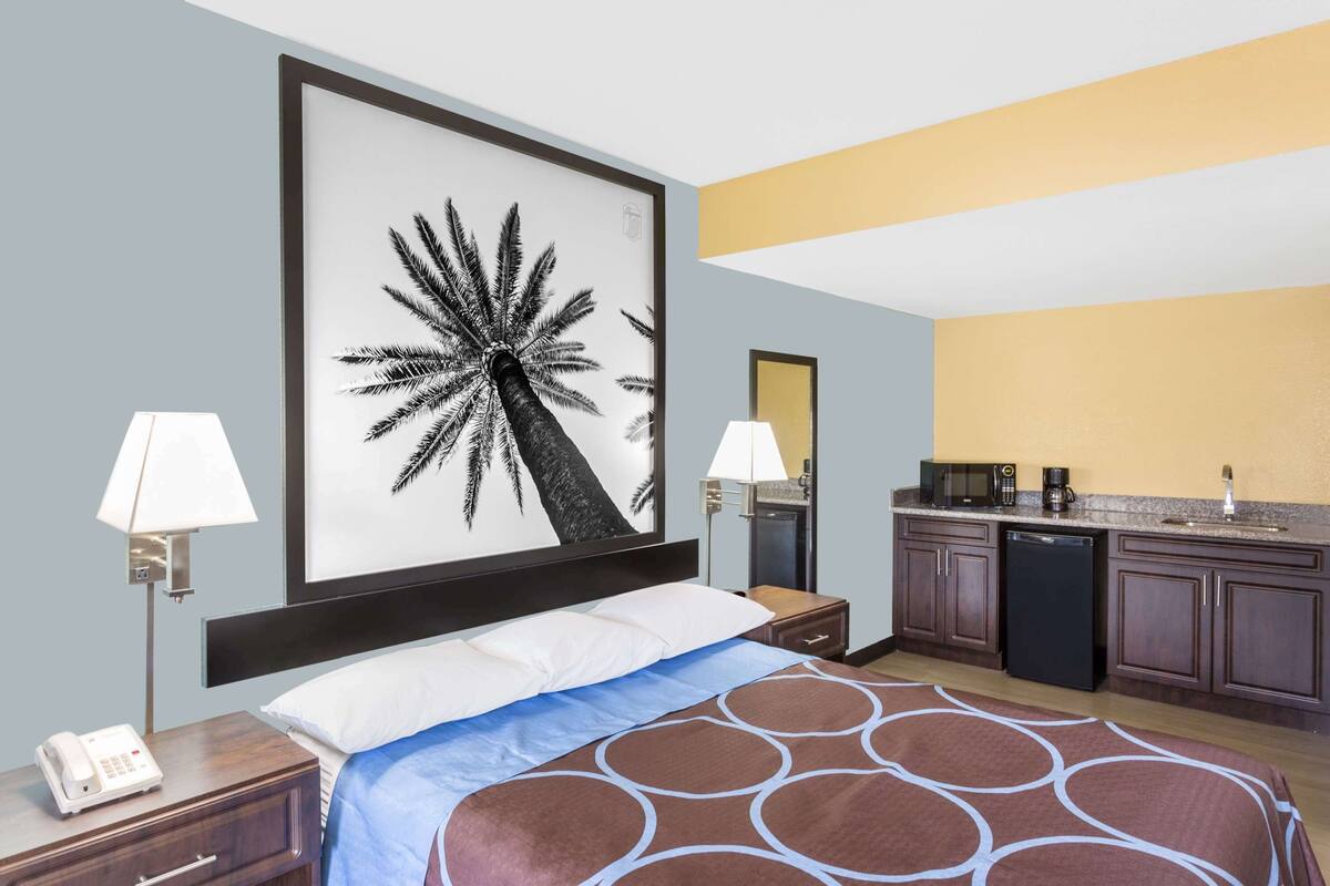 Une chambre d’hôtel comprenant un lit, des lampes de chevet, un grand tableau encadré, une kitchenette et une table de chevet avec un téléphone.
