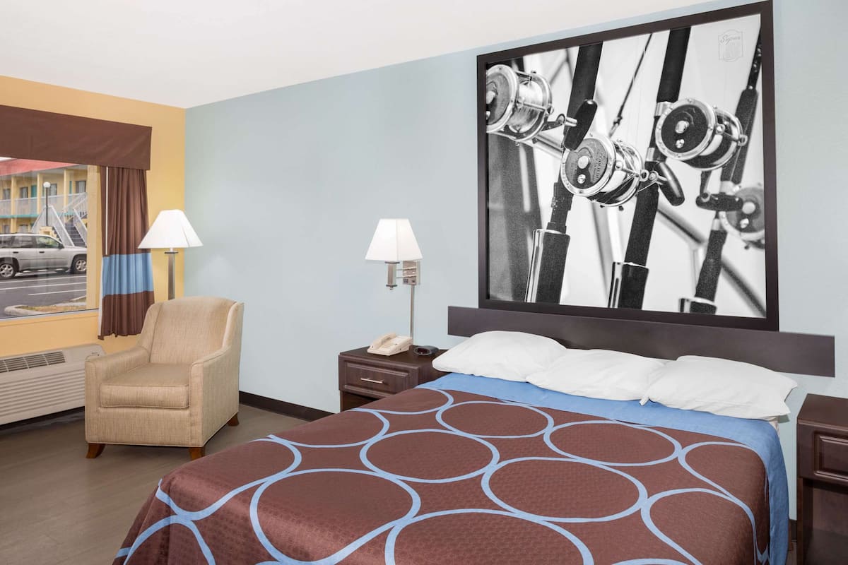 Une chambre d’hôtel avec un lit, une chaise, une table de chevet, une lampe et un grand tableau encadré au mur.