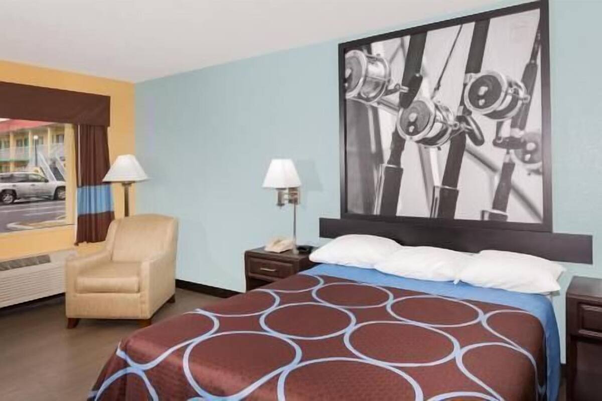 Une chambre d’hôtel avec un lit, une chaise, une table de chevet, une lampe et un mur orné d’un grand tableau abstrait.