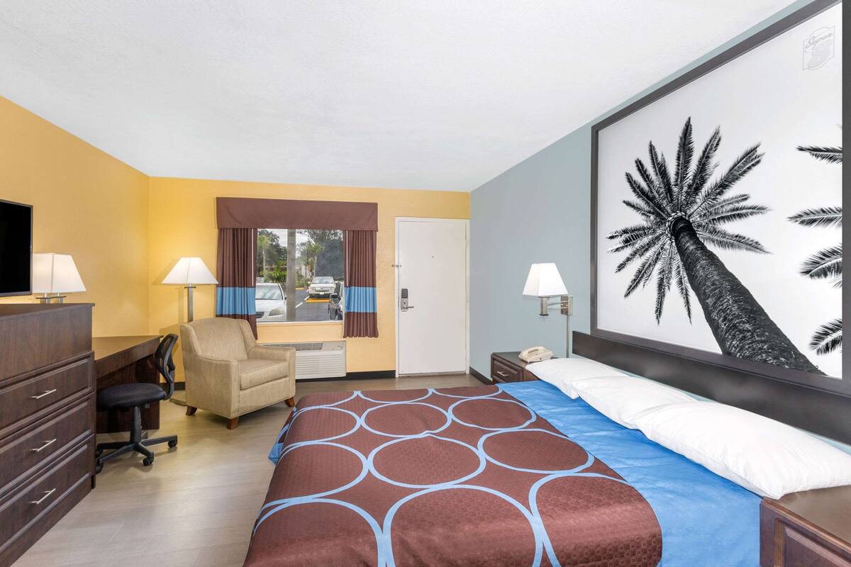 Une chambre d’hôtel avec un lit, un bureau, une chaise et un grand tableau représentant un palmier.