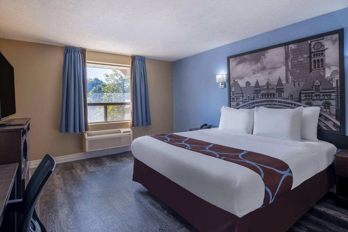 Ein Hotelzimmer mit einem großen Bett, einem Schreibtisch mit Computer, einem Fenster mit Vorhängen und einem Wandbild.