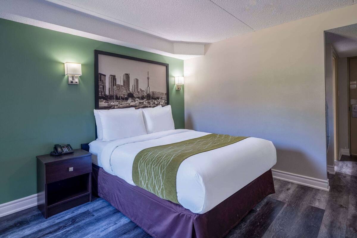Ein Hotelzimmer mit Bett, Nachttisch, Stadtbild-Kunstwerk und einer Tür zu einem weiteren Raum.
