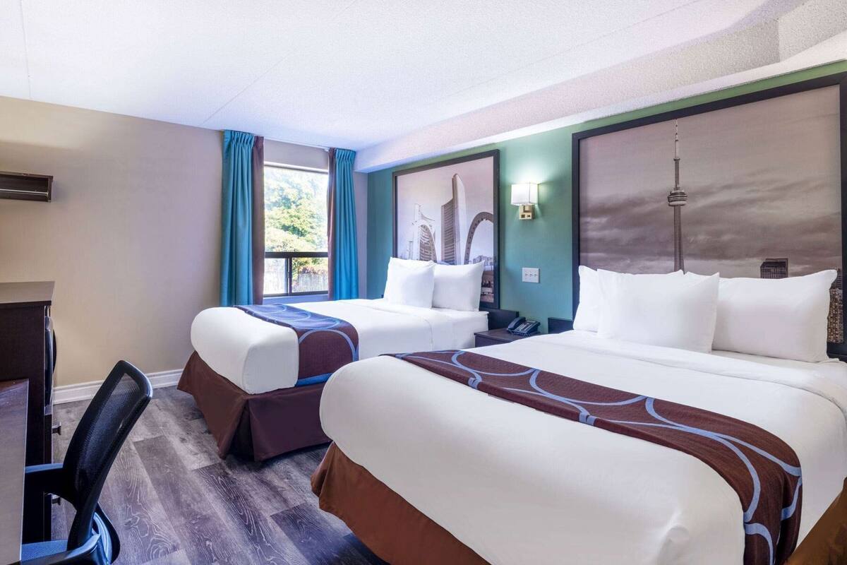 Ein Hotelzimmer mit zwei Betten, einem Schreibtisch, einem Stuhl, einem Fenster mit Vorhängen, einer Wandlampe und Blick auf die Stadt.