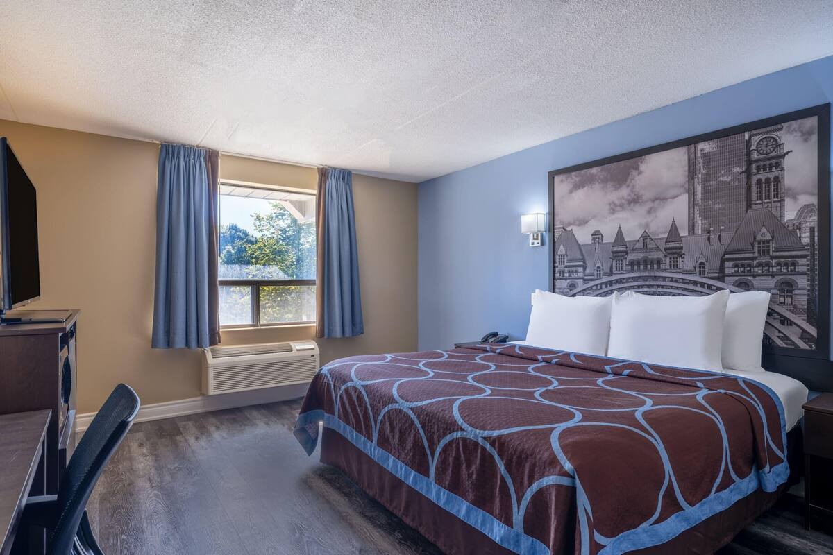 Ein Hotelzimmer mit einem großen Bett, einem Schreibtisch, einem Stuhl, einem Fenster mit Vorhängen und einem Wandgemälde mit Stadtansicht.