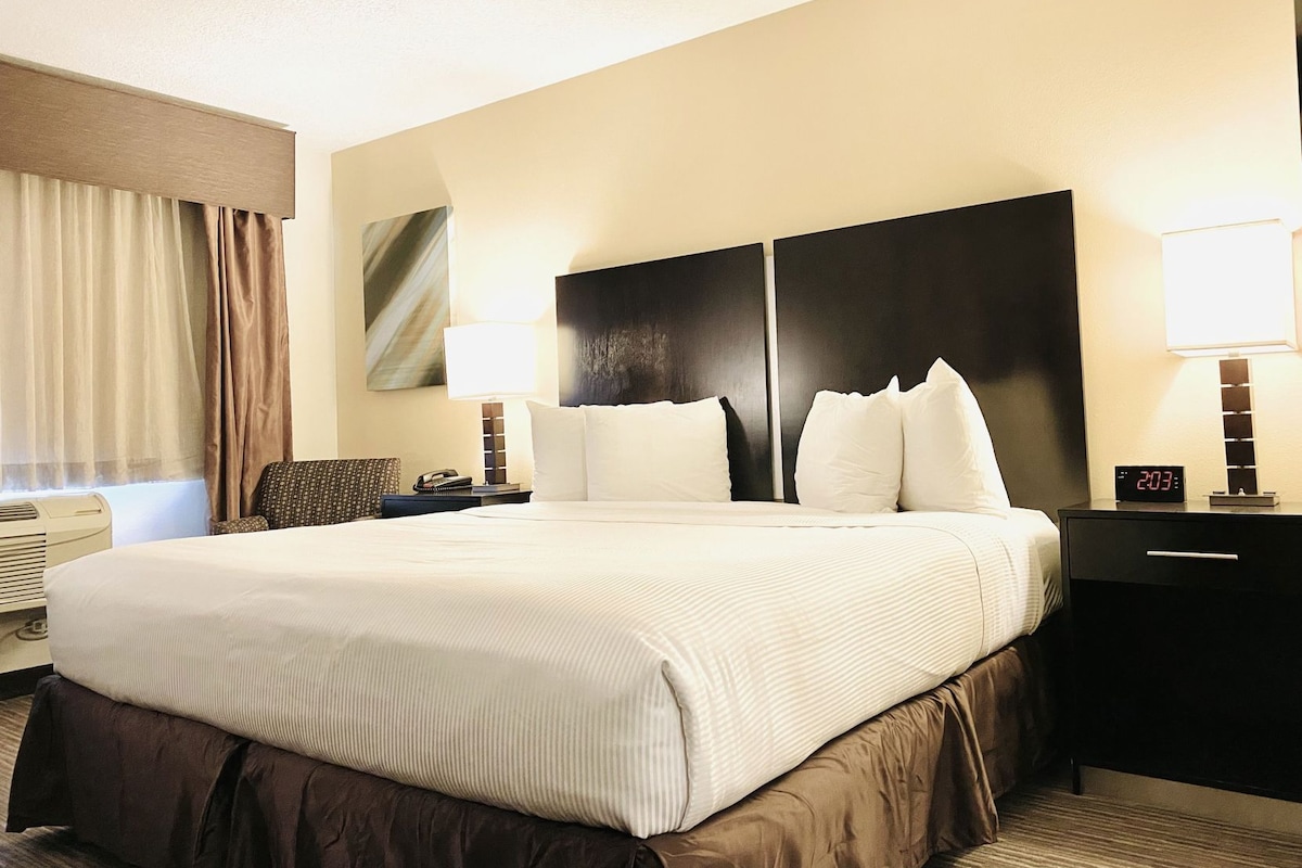 A hotel room with a large bed, two bedside tables, a nightstand, a lamp, a clock, and a window with curtains.