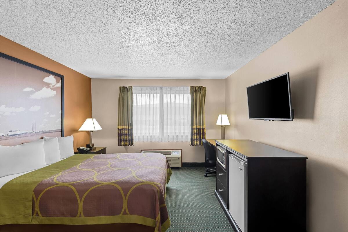 Habitación de hotel con cama, escritorio, televisor y un amplio ventanal con cortinas.